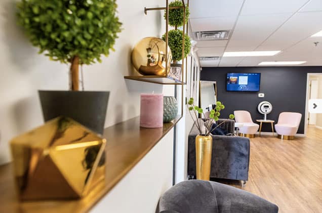 A soothing and beautiful patient waiting area with comfortable and soft seating, hardwood floors, gold accents, green plants, and a hardwood floor in KP Aesthetic waiting area.