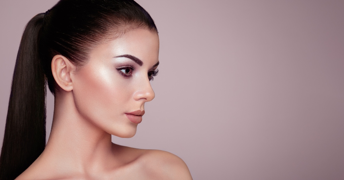 A young woman with flawless, glowing skin and sleek, dark hair pulled back into a ponytail is shown in profile against a soft mauve background