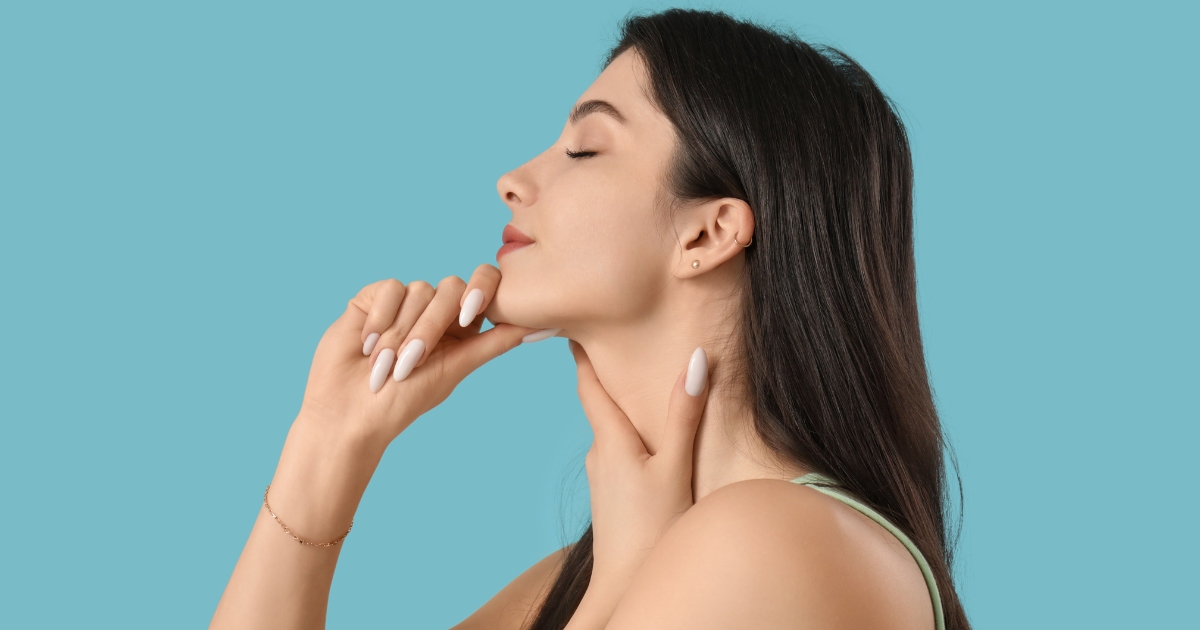 A person with long dark hair is shown in profile against a solid light blue background, gently touching their jawline and neck with their hands while keeping their eyes closed.